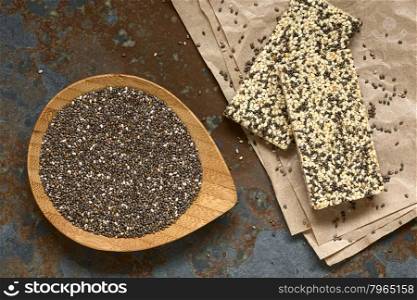 Chia seeds (lat. Salvia hispanica) on small bamboo plate with chia-sesame-honey granola bar on the side, photographed overhead on slate with natural light. Chia seeds are considered a superfood containing protein, omega fat, minerals, antioxidants.