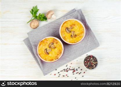 Chicken and mushroom casserole with golden crust, julienne with baked cheese, creamy gratin in portion ramekin clay pot on white wooden background. French cuisine. Top view, flat lay. French Julienne casserole with chicken and mushroom in portion ramekin on white background