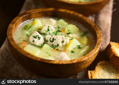 Chicken and potato chowder soup with green bell pepper and carrot in wooden bowl garnished with fresh parsley, photographed with natural light (Selective Focus, Focus in the middle of the soup)
