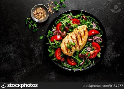 Chicken breast fillet grilled and fresh vegetable green salad with arugula, tomatoes and olives on black background, healthy food, mediterranean diet, top view
