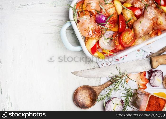 Chicken breast with cut colorful vegetables in enamelled casserole with wooden spoon and kitchen knife on white wooden background, top view, border. Healthy food or diet nutrition concept.