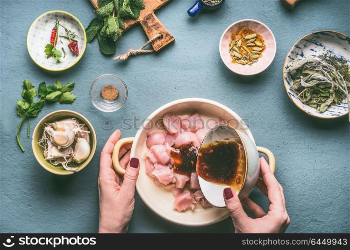 Chicken meal cooking concept. Female hands marinates chicken pieces in bowl with oil and soy sauce on kitchen tables background with ingredients, top view