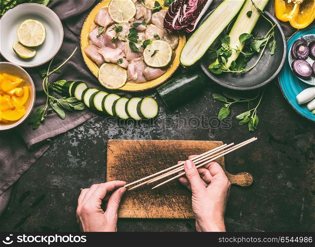 Chicken meat and vegetables skewers making. Female hands holding wooden skewer on kitchen table background with chicken pieces and vegetables, top view. Grill preparation