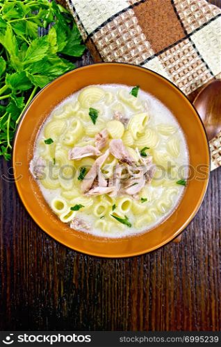 Chicken meat soup, pasta with cream and cilantro in a clay plate, towel, parsley, spoon on a wooden board background from above