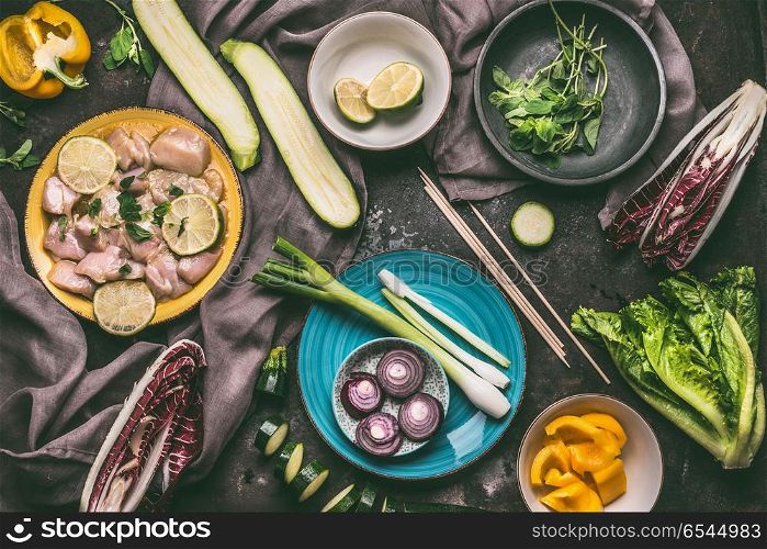 Chicken meat with lemon and vegetables skewers. Grill preparation on kitchen table background with chicken pieces and vegetables in plates and bowls, top view.