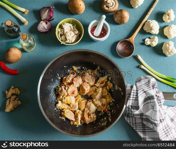 Chicken pieces with spices in wok frying pan on kitchen table with vegetables ingredients and wooden spoon, top view. Cooking preparation