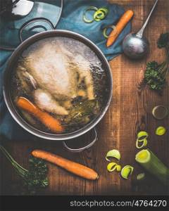 Chicken soup cooking, pot with chicken broth and ladle on dark rustic wooden background with vegetables ingredients, top view. Healthy food or Diet nutrition concept