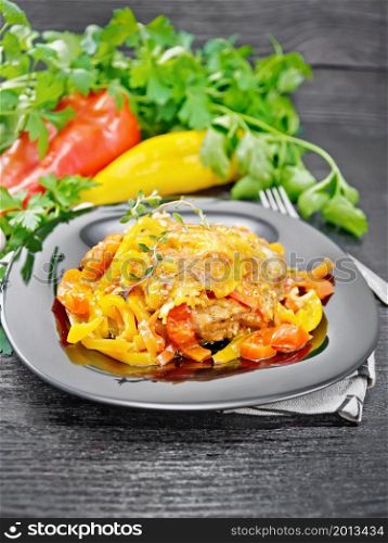 Chicken stewed with tomatoes, yellow and red bell peppers and cheese in a plate on napkin, thyme, parsley and garlic on black wooden board background