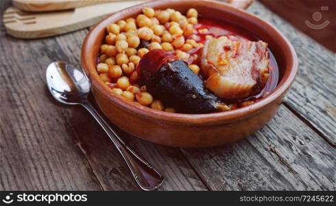 Chickpeas, sausage and bacon in a crockpot by wooden spoon and fork, and a little metal spoon. Typical food from Madrid, Spain, with a rustic wooden board as a background.