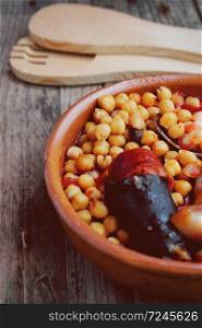 Chickpeas, sausage and bacon in a crockpot by wooden spoon and fork. Typical food from Madrid, Spain, with a rustic wooden board as a background.