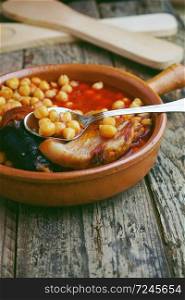 Chickpeas, sausage and bacon in a crockpot by wooden spoon and fork, and a little metal spoon. Typical food from Madrid, Spain, with a rustic wooden board as a background.