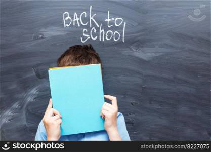 child boy covered his face with a textbook near a chalkboard. Back to school concept background. child boy covered his face with a textbook near chalkboard. Back to school concept background