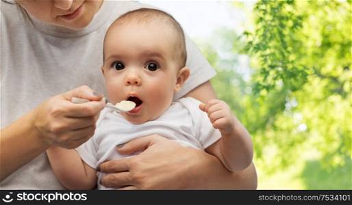 child, eating and food concept - close up of mother with spoon feeding little baby over green natural background. close up of mother with spoon feeding little baby