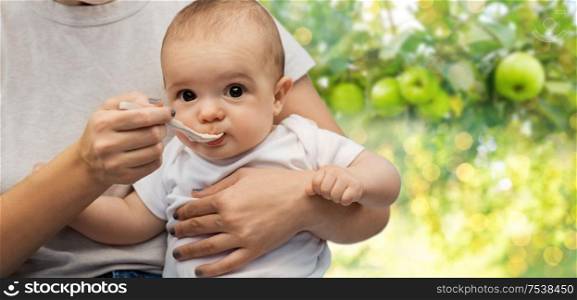 child, eating and food concept - close up of mother with spoon feeding little baby over green apples on summer garden background. close up of mother with spoon feeding little baby