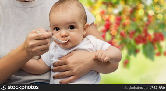 child, eating and food concept - close up of mother with spoon feeding little baby over green natural background. close up of mother with spoon feeding little baby