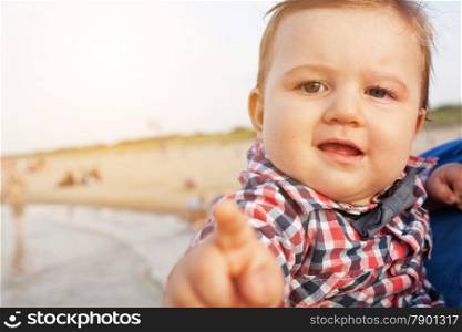 Child pointing at the camera with funny expression being hold by a father on the beach.