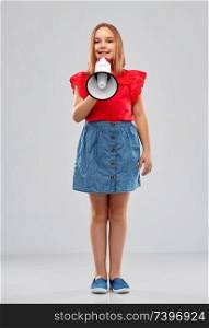 child rights, communication and people concept - smiling girl in red shirt and denim skirt speaking to megaphone over grey background. smiling girl speaking to megaphone