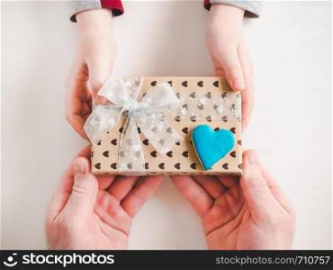 Child's hands and adult man's hands, beautiful gift box, ribbon and glazed cookies on a white, wooden background. Top view, close-up. Preparing for the holidays. Beautiful card for Women's Equality Day. Close-up