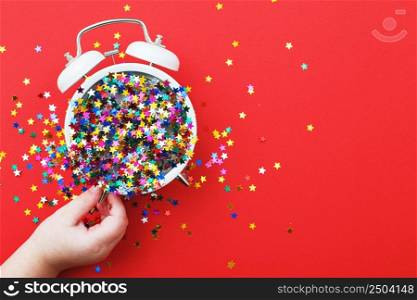 child&rsquo;s hands hold a white alarm clock with scattered sequins in the form of stars on a red background. Party, christmas, new year, celebration concept. copy space. Flat lay. Top view. child&rsquo;s hands hold alarm clock with scattered sequins on a red background. christmas, celebration concept