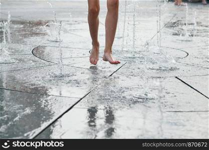 Child standing, jumping, playing in a fountain while hot weather. Closeup of legs, water streams, splashes. Candid people, real moments, authentic situations