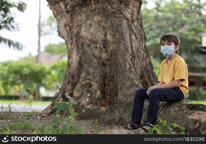Child wearing face mask sitting under big tree and looking at camera.Pollution concept.