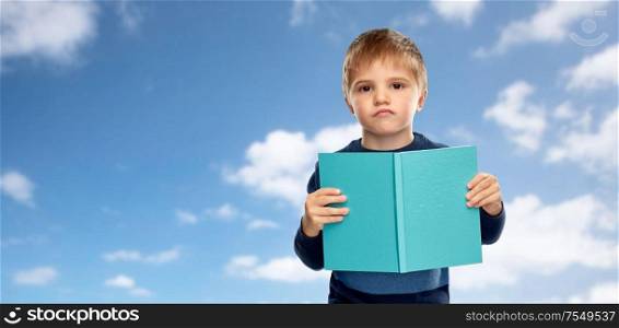 childhood and education concept - displeased little boy reading book over blue sky and clouds background. displeased little boy with book over blue sky
