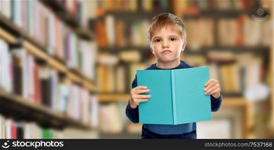 childhood and education concept - displeased little boy reading book over library background. displeased little boy reading book over library