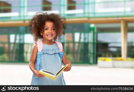 childhood and education concept - happy little african american girl with book and backpack over school yard background. happy little african girl with book and backpack