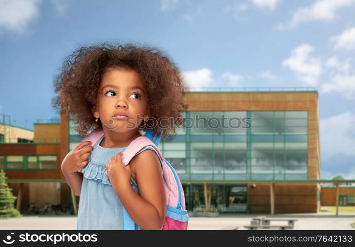 childhood and education concept - sad little african american girl with backpack over school on background. sad little african american girl with backpack