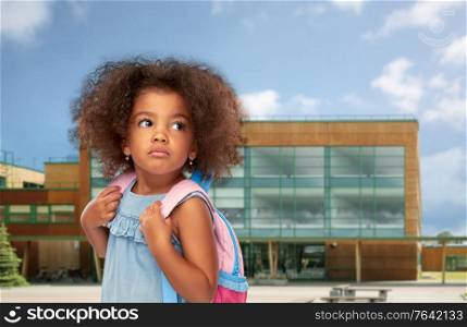 childhood and education concept - sad little african american girl with backpack over school on background. sad little african american girl with backpack