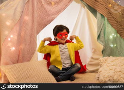 childhood and hygge concept - happy little boy wearing superhero mask and cape in kids tent or teepee at home. happy boy in super hero stuff in kids tent at home. happy boy in super hero stuff in kids tent at home