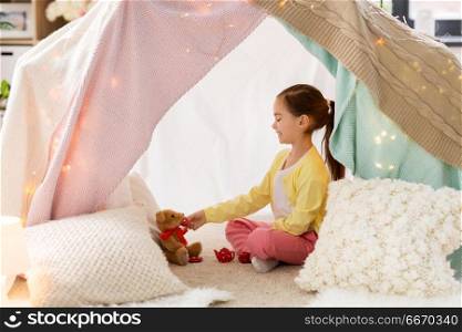 childhood and hygge concept - happy little girl playing tea party with teddy bear in kids tent at home. little girl playing tea party in kids tent at home. little girl playing tea party in kids tent at home