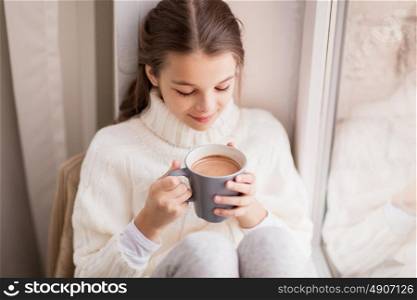 childhood and people concept - beautiful girl with cacao mug in sweater sitting at home window in winter. girl with cacao mug sitting at home window