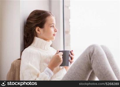 childhood and people concept - beautiful girl with tea mug in sweater sitting at home window in winter. girl with tea mug sitting at home window