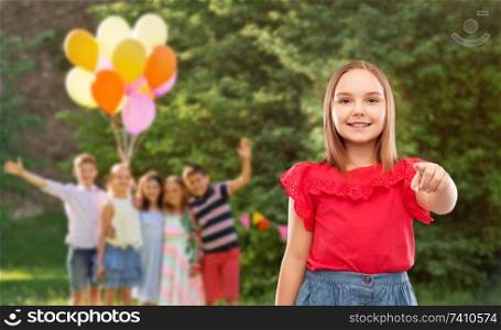 childhood and people concept - beautiful smiling girl in red shirt and skirt pointing to you at birthday party over friends in summer park background. smiling girl pointing to you at birthday party