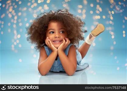childhood and people concept - happy little african american girl lying on floor over festive lights on blue background. happy little african american girl lying on floor