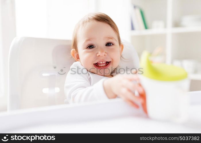 childhood and people concept - happy smiling little baby drinking from spout cup sitting in highchair at home. baby drinking from spout cup in highchair at home