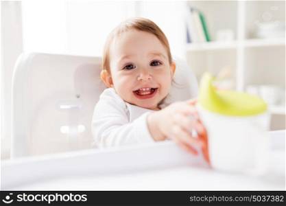 childhood and people concept - happy smiling little baby drinking from spout cup sitting in highchair at home. baby drinking from spout cup in highchair at home