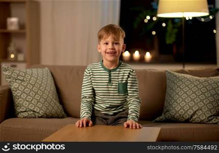 childhood and people concept - happy smiling little boy sitting in sofa at home at night. happy smiling little boy at home at night