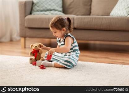 childhood and people concept - happy three years old baby girl playing tea party with toy crockery and teddy bear at home. little girl playing with toy tea set at home