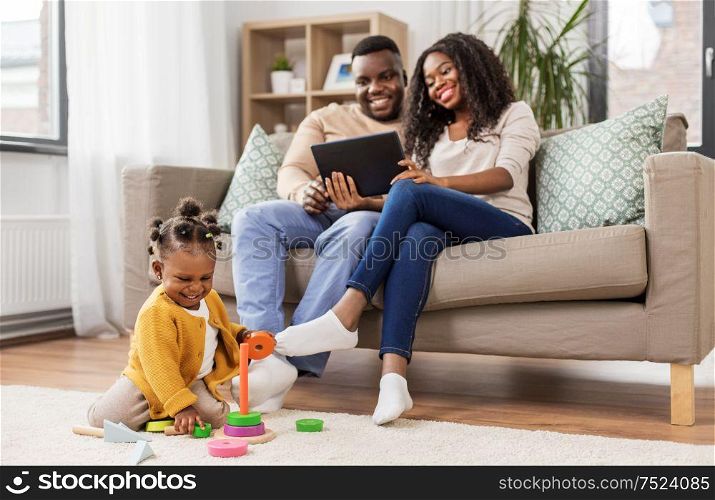 childhood and people concept - little african american baby girl playing with toy blocks and parents with tablet pc computer at home. african baby girl playing with toy blocks at home