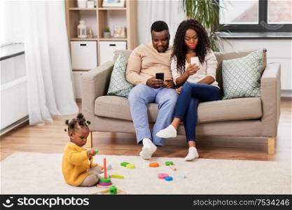 childhood and people concept - little african american baby girl playing with toy blocks and parents using smartphones at home. african baby girl playing with toy blocks at home