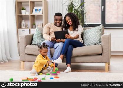 childhood and people concept - little african american baby girl playing with toy blocks and parents with tablet pc computer at home. family with tablet pc and toy blocks at home