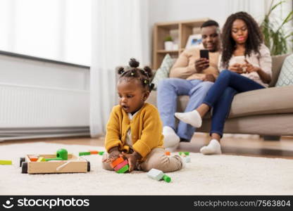 childhood and people concept - little african american baby girl playing with toy blocks and parents using smartphones at home. african baby girl playing with toy blocks at home