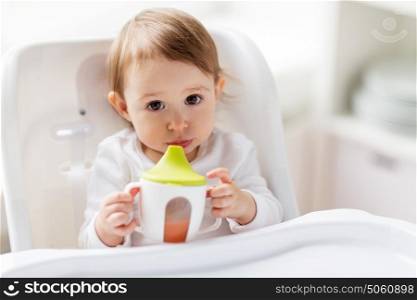 childhood and people concept - little baby drinking from spout cup sitting in highchair at home. baby drinking from spout cup in highchair at home