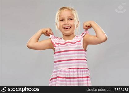 childhood and people concept - smiling little girl in striped dress showing her power over grey background. smiling little girl showing her power