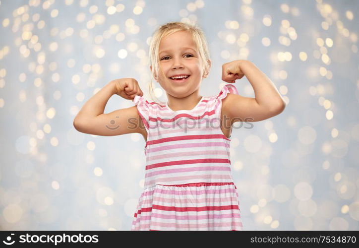 childhood and people concept - smiling little girl in striped dress showing her power over festive lights background. smiling little girl showing her power
