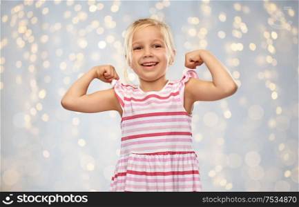 childhood and people concept - smiling little girl in striped dress showing her power over festive lights background. smiling little girl showing her power