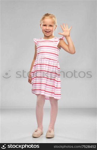 childhood and people concept - smiling little girl in striped dress waving hand over grey background. smiling little girl in striped dress