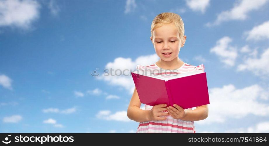 childhood and people concept - smiling little girl reading book over blue sky and clouds background. smiling little girl reading book